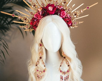Puede incluir: Una tiara dorada con puntas radiantes, adornada con rosas rojas y rosas y piedras preciosas. Pendientes colgantes a juego con cuentas rosas colgantes. La tiara y los pendientes se exhiben en una cabeza de maniquí blanca con pelo rubio largo y ondulado.