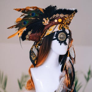 Könnte beinhalten: Ein dekorativer Kopfschmuck mit einem Farbton aus Schwarz, Braun und Gold. Der Kopfschmuck verfügt über Pfauenfedern, goldene Verzierungen und eine schwarze Spitzen-Augenmaske. Zusätzliche Federn fallen an der Seite herab und erzeugen einen dramatischen und verzierten Look.