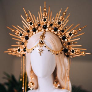May include: A gold-toned headpiece with a sunburst design, adorned with black and gold accents. The headpiece features a central pendant and dangling chains. The design is displayed on a white mannequin head, suggesting a costume or accessory.