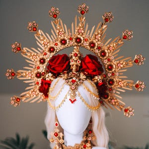 Pode incluir: Uma tiara dourada com detalhes vermelhos e rosas, exibida em uma cabeça de manequim branca. A tiara apresenta um design de raios de sol, detalhes florais intrincados e correntes penduradas. Brincos e colar combinando completam o conjunto.