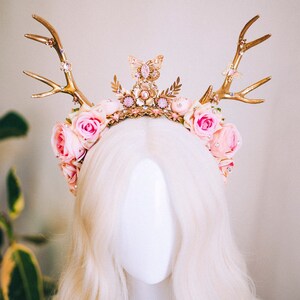 Puede incluir: Una diadema de asta de ciervo dorada adornada con flores rosas y un dije de mariposa. La diadema est&aacute; decorada con diamantes de imitaci&oacute;n y cuentas.