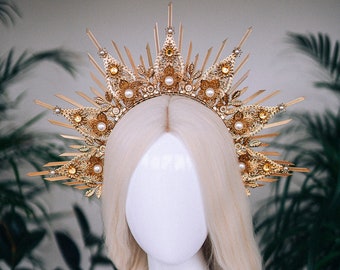 May include: A gold crown with a sunburst design and pearl accents. The crown is on a white mannequin head with blonde hair.
