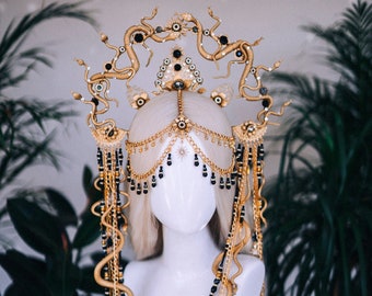 May include: A gold and black headpiece with a snake design. The headpiece is made of metal and beads and has a crown-like shape. It is worn on a white mannequin head.