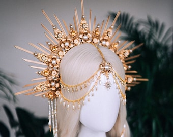 May include: A gold-colored headpiece with a sunburst design, adorned with pearls and floral accents. The headpiece features dangling chains and star-shaped charms. The item is displayed on a white mannequin head with blonde hair.