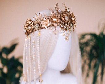 May include: A gold crown with a moon and star design, featuring white feathers and beads hanging from the bottom. The crown is on a white mannequin head.