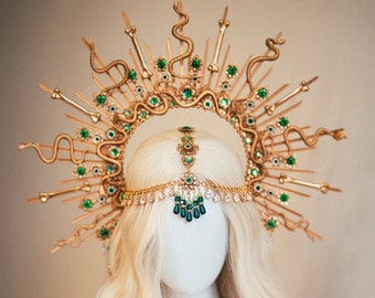 May include: A gold crown with green gemstones and snake details. The crown is on a white mannequin head with long blonde hair.