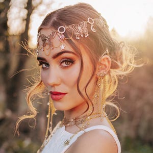 May include: A woman wearing a white lace dress and gold jewelry, including a headpiece, ear cuffs, and layered necklaces. The jewelry has celestial and butterfly motifs. The background is a blurred outdoor scene with sunlight.
