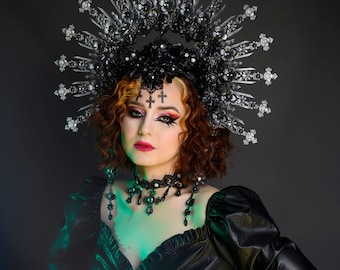 May include: A woman wearing a black leather top with a large silver and black crown with crosses. She has dark makeup with red lips and is wearing a black choker necklace with dangling crystals.