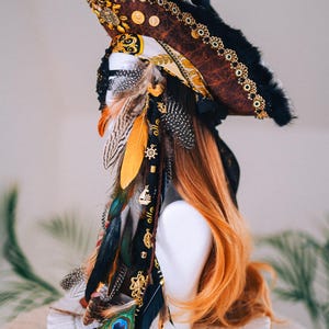 Puede incluir: Un sombrero de pirata decorativo con un acabado marr&oacute;n similar al cuero, ribete negro y detalles dorados. El sombrero est&aacute; adornado con plumas de colores, abalorios dorados y una pluma de pavo real. El sombrero se exhibe sobre una cabeza de maniqu&iacute; blanca con pelo largo y casta&ntilde;o rojizo.