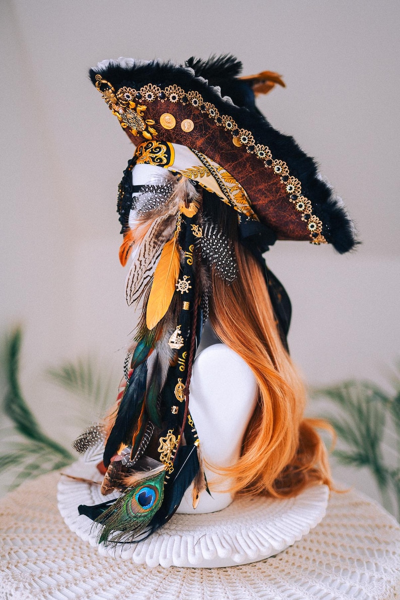 Puede incluir: Un sombrero de pirata decorativo con un acabado marr&oacute;n similar al cuero, ribete negro y detalles dorados. El sombrero est&aacute; adornado con plumas de colores, abalorios dorados y una pluma de pavo real. El sombrero se exhibe sobre una cabeza de maniqu&iacute; blanca con pelo largo y casta&ntilde;o rojizo.