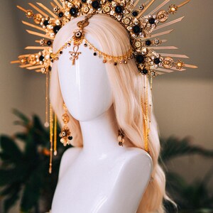May include: A gold-toned headpiece with a sunburst design, adorned with black gemstones and intricate details. The headpiece is displayed on a white mannequin head with blonde hair, showcasing its ornate craftsmanship and celestial theme.