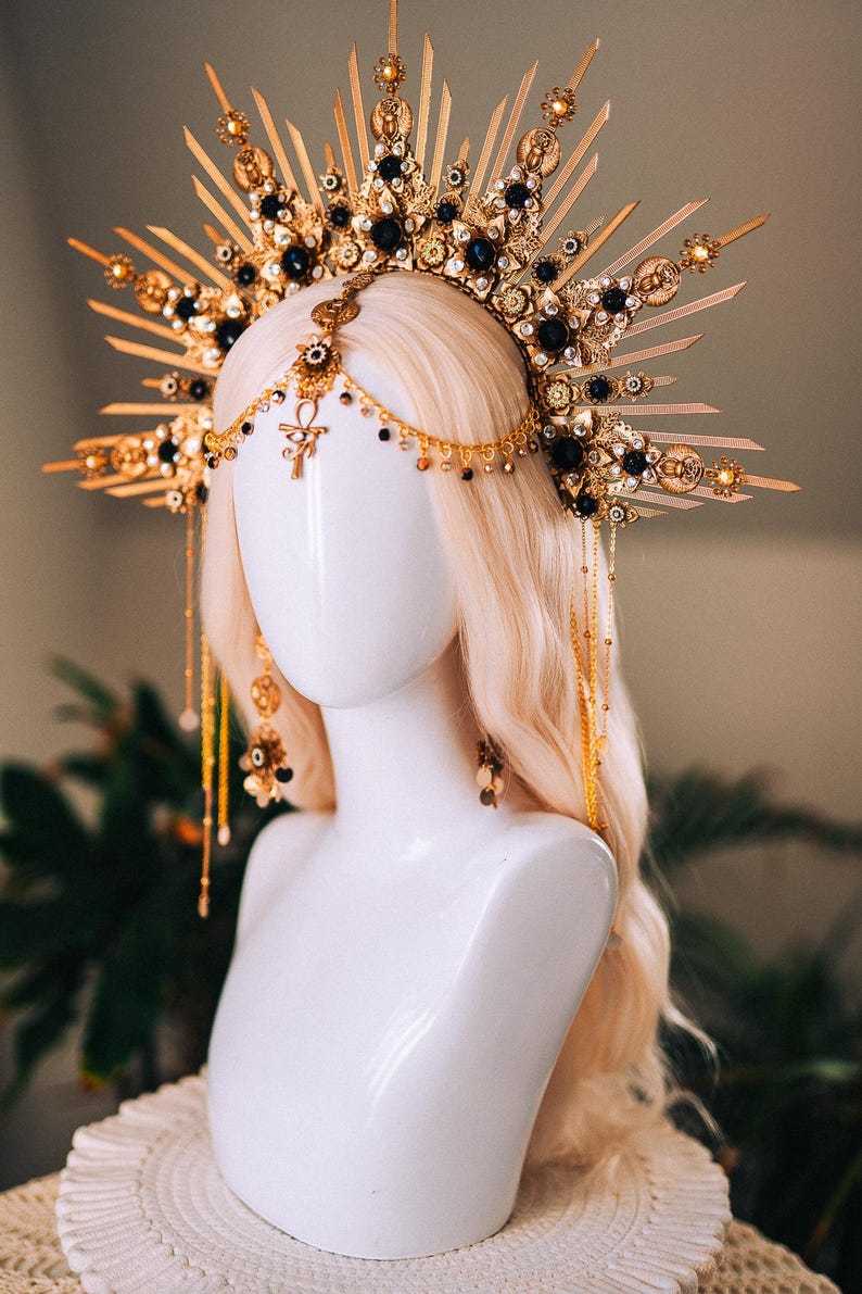 May include: A gold-toned headpiece with a sunburst design, adorned with black gemstones and intricate details. The headpiece is displayed on a white mannequin head with blonde hair, showcasing its ornate craftsmanship and celestial theme.