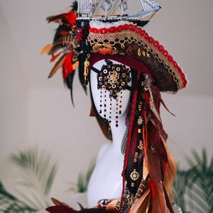 以下が含まれることがあります： ミニチュア船が飾られた海賊帽子。赤、茶色、金色の羽が特徴で、赤と金のレーストリム、装飾が施された黒い眼帯、ぶら下がっているビーズが付いています。全体的なデザインは複雑で、演劇的です。