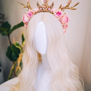 Puede incluir: Una diadema de asta de ciervo dorada con flores rosas y un dije de mariposa. La diadema est&aacute; en una cabeza de maniqu&iacute; blanca con cabello largo y rubio.