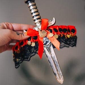 以下が含まれることがあります： 赤いリボン、黒いレース、金色のアクセントが付いた装飾用の短剣。短剣は金色の柄と銀色の刃が特徴です。スカルとクロスボーンの小さな金色のチャームがリボンに付いています。コスチュームアクセサリーです。