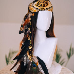 Puede incluir: Tocado decorativo con un pa&ntilde;uelo de seda estampado en oro y negro, adornado con plumas en tonos marr&oacute;n, negro y naranja. A las plumas se les adjuntan abalorios dorados. El tocado se exhibe sobre una cabeza de maniqu&iacute; blanca.