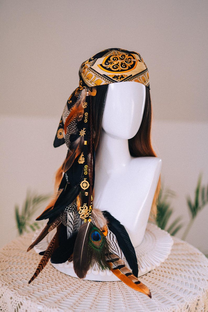 Puede incluir: Tocado decorativo con un pa&ntilde;uelo de seda estampado en oro y negro, adornado con plumas en tonos marr&oacute;n, negro y naranja. A las plumas se les adjuntan abalorios dorados. El tocado se exhibe sobre una cabeza de maniqu&iacute; blanca.
