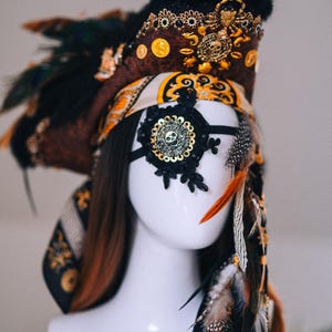 Puede incluir: Tocado elaborado con una m&aacute;scara para los ojos con tem&aacute;tica de calavera. El tocado presenta detalles en marr&oacute;n, negro y dorado, incluyendo plumas, monedas y adornos ornamentados. La m&aacute;scara para los ojos es de encaje negro con un dise&ntilde;o de calavera dorada.
