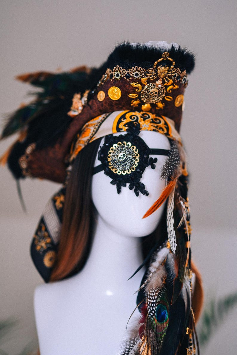 Puede incluir: Tocado elaborado con una m&aacute;scara para los ojos con tem&aacute;tica de calavera. El tocado presenta detalles en marr&oacute;n, negro y dorado, incluyendo plumas, monedas y adornos ornamentados. La m&aacute;scara para los ojos es de encaje negro con un dise&ntilde;o de calavera dorada.