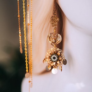 May include: Gold-toned statement earrings with an Egyptian theme. The earrings feature a scarab beetle, a snake, and a floral design with a black center stone. Delicate chains and small circular charms dangle from the earrings, creating movement and visual interest.