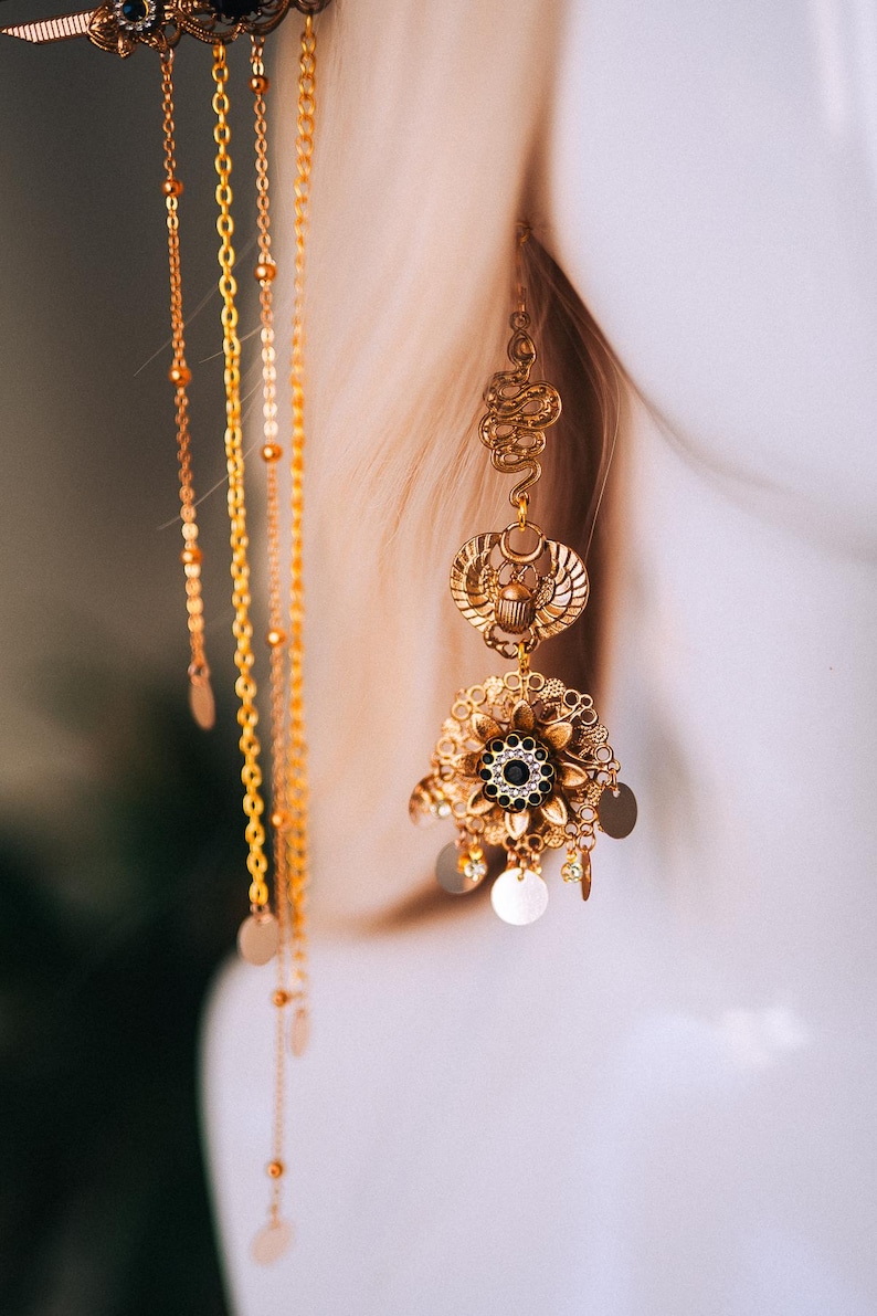 May include: Gold-toned statement earrings with an Egyptian theme. The earrings feature a scarab beetle, a snake, and a floral design with a black center stone. Delicate chains and small circular charms dangle from the earrings, creating movement and visual interest.