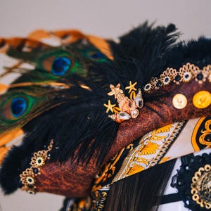 Puede incluir: Un tocado marr&oacute;n y negro con plumas de pavo real, plumas negras y adornos dorados. El tocado presenta un broche de conchas marinas y estrellas de mar, monedas de oro y una bufanda estampada.