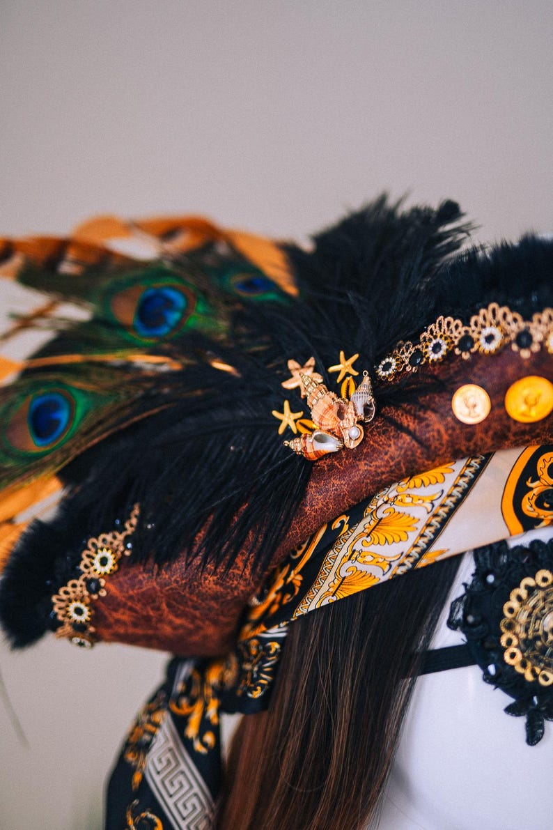 Puede incluir: Un tocado marr&oacute;n y negro con plumas de pavo real, plumas negras y adornos dorados. El tocado presenta un broche de conchas marinas y estrellas de mar, monedas de oro y una bufanda estampada.