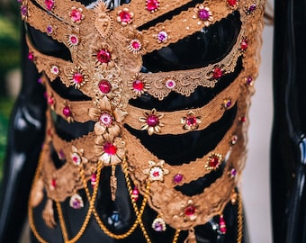 Könnte beinhalten: Ein goldfarbenes, verziertes Mieder mit rosa und roten Juwelen. Das Mieder ist aus Spitze und hat ein zartes, weibliches Design.