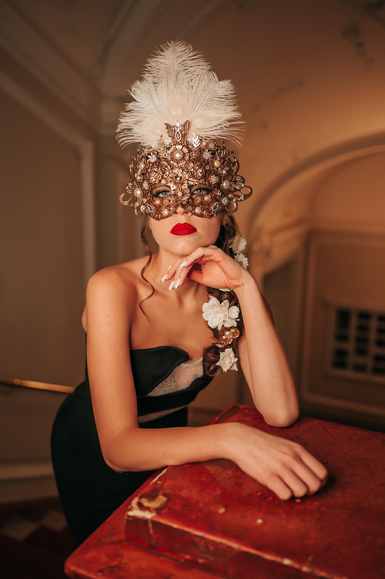 K&ouml;nnte beinhalten: Eine Frau tr&auml;gt eine goldene und wei&szlig;e Maskenballmaske mit Federn und Strasssteinen. Sie tr&auml;gt ein schwarzes tr&auml;gerloses Kleid und hat roten Lippenstift auf. Sie lehnt sich an eine rote Holzoberfl&auml;che.