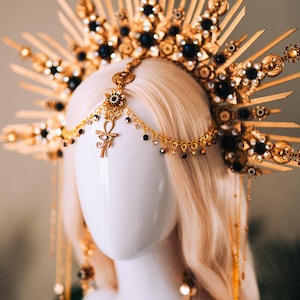 May include: A gold-toned headpiece with a sunburst design, adorned with black gemstones and intricate details. A chain with an Ankh pendant and dangling beads hangs from the center. The headpiece is displayed on a mannequin head with blonde hair.