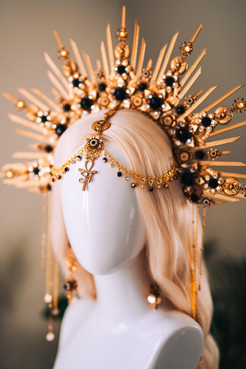 May include: A gold-toned headpiece with a sunburst design, adorned with black gemstones and intricate details. A chain with an Ankh pendant and dangling beads hangs from the center. The headpiece is displayed on a mannequin head with blonde hair.