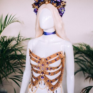May include: A gold and purple skeleton-inspired corset with a skull and flower crown. The corset is made of lace and features a variety of embellishments, including rhinestones and chains.