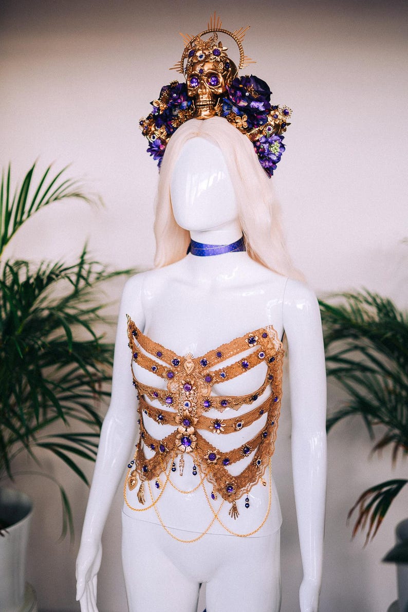 May include: A gold and purple skeleton-inspired corset with a skull and flower crown. The corset is made of lace and features a variety of embellishments, including rhinestones and chains.