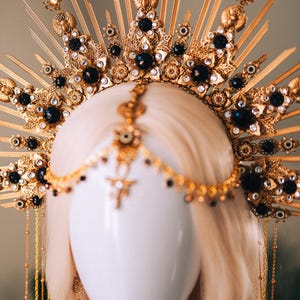 May include: A gold-colored headpiece with intricate details, featuring black gemstones and decorative elements. The headpiece has a sunburst design with dangling chains and is displayed on a mannequin head.