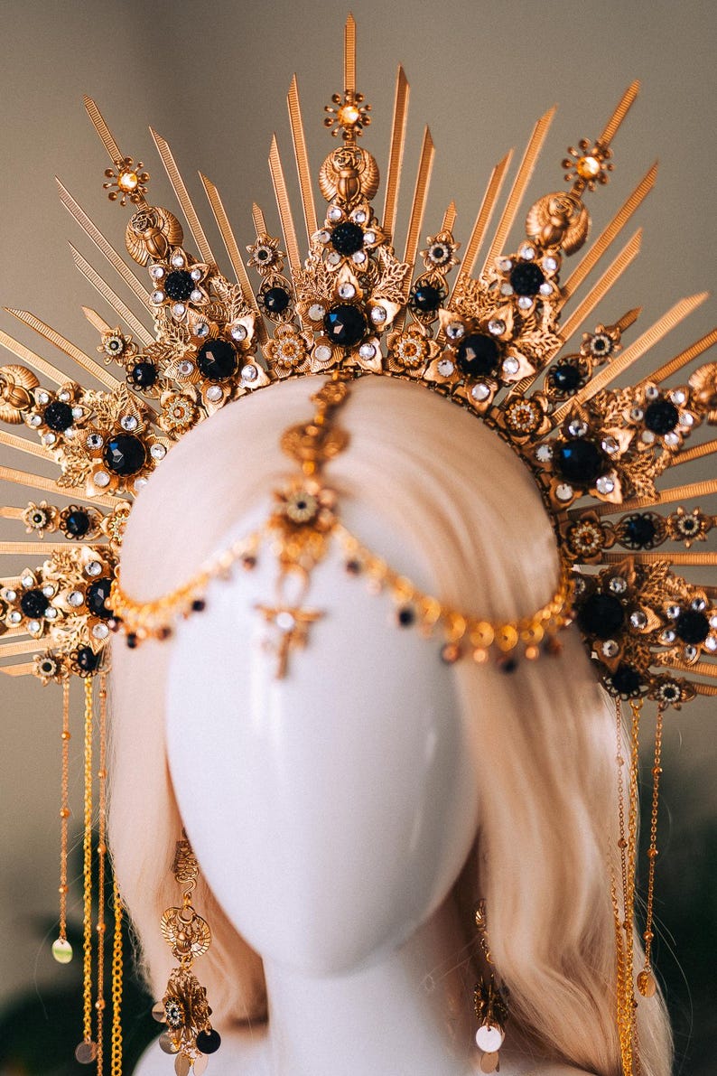May include: A gold-colored headpiece with intricate details, featuring black gemstones and decorative elements. The headpiece has a sunburst design with dangling chains and is displayed on a mannequin head.
