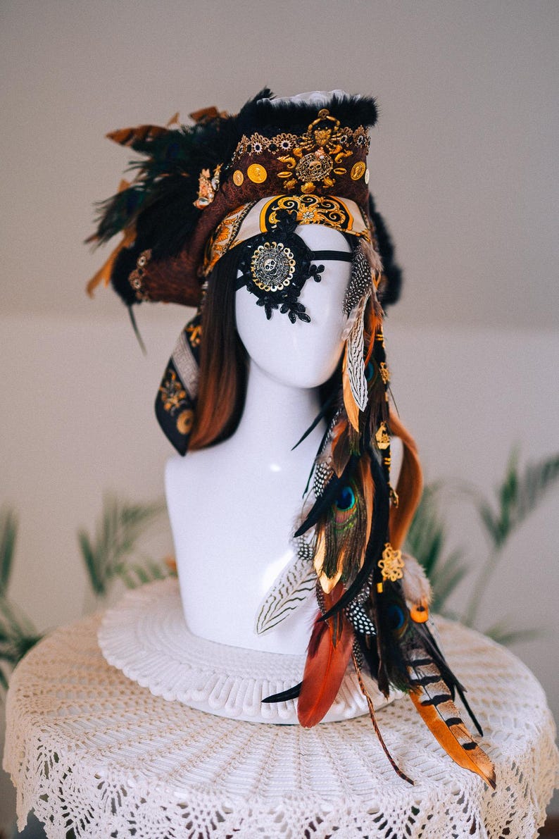 Puede incluir: Tocado decorativo con una mezcla de plumas marrones, naranjas y negras. El tocado incluye una m&aacute;scara de ojos negra con adornos dorados y un adorno decorativo para la cabeza con detalles dorados. El art&iacute;culo se exhibe en una cabeza de maniqu&iacute; blanca.