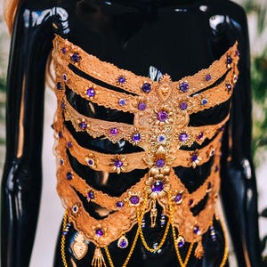 May include: A gold and purple embellished corset with a lace design and dangling chains. The corset is on a black mannequin.