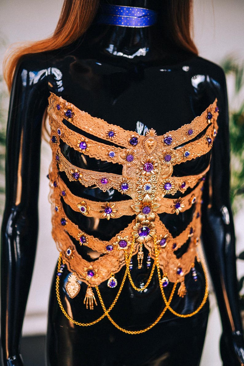 May include: A gold and purple embellished corset with a lace design and dangling chains. The corset is on a black mannequin.