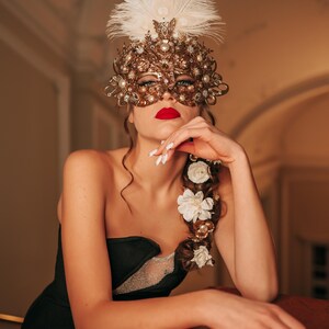 K&ouml;nnte beinhalten: Eine Frau tr&auml;gt eine goldene und wei&szlig;e Maskenballmaske mit Federn und ein schwarzes tr&auml;gerloses Kleid. Sie hat wei&szlig;e Blumen im Haar und posiert mit der Hand am Kinn.