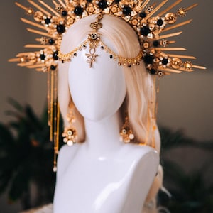 May include: A gold-toned headpiece with a sunburst design, adorned with black beads and intricate detailing. The headpiece is displayed on a white mannequin head with blonde hair. The design includes dangling chains and an Ankh symbol.