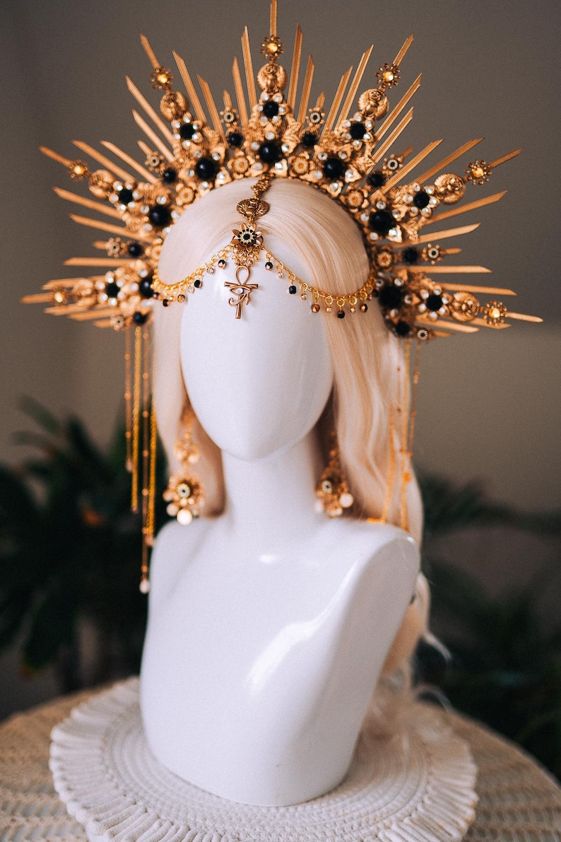 May include: A gold-toned headpiece with a sunburst design, adorned with black beads and intricate detailing. The headpiece is displayed on a white mannequin head with blonde hair. The design includes dangling chains and an Ankh symbol.