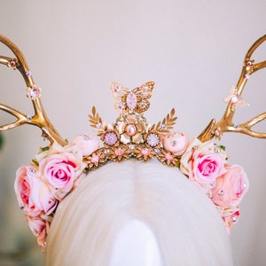 Puede incluir: Una diadema de asta de ciervo dorada adornada con rosas rosas y un dije de mariposa. La diadema est&aacute; decorada con diamantes de imitaci&oacute;n y otros adornos.