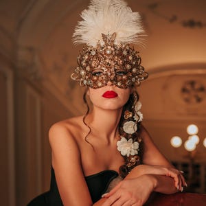 K&ouml;nnte beinhalten: Eine Frau tr&auml;gt eine goldene und wei&szlig;e Maskenballmaske mit Federn und Strasssteinen. Sie tr&auml;gt ein schwarzes tr&auml;gerloses Kleid und hat roten Lippenstift.