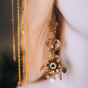 May include: Close-up of an ornate gold earring with multiple layers and dangling chains. The earring features a scarab beetle design, floral elements, and small circular charms. The style is reminiscent of ancient Egyptian jewelry.