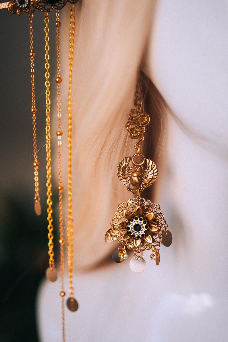 May include: Close-up of an ornate gold earring with multiple layers and dangling chains. The earring features a scarab beetle design, floral elements, and small circular charms. The style is reminiscent of ancient Egyptian jewelry.