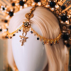 May include: A gold-toned headpiece with an elaborate design, featuring a sunburst crown and dangling chains. The headpiece includes black and clear gemstones and an ankh pendant. The headpiece is displayed on a white mannequin head with blonde hair.