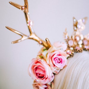 Puede incluir: Una diadema de asta de ciervo dorada con rosas rosas y pedrer&iacute;a. La diadema se lleva en una persona con cabello rubio.