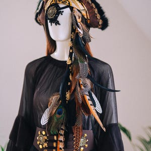 Puede incluir: Un sombrero pirata marr&oacute;n con adornos dorados, ribete de piel negra y una bufanda decorativa. El sombrero est&aacute; adornado con plumas de varios colores, incluyendo pavo real, naranja y negro. Una m&aacute;scara de ojos negra y un cintur&oacute;n marr&oacute;n con monedas de oro completan el conjunto.