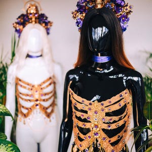 May include: Two mannequins wearing elaborate gold and purple embellished tops. The tops are designed to resemble a ribcage and feature a variety of gems and beads. The mannequins are wearing elaborate headpieces with skulls and flowers.