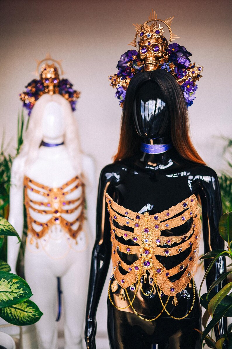 May include: Two mannequins wearing elaborate gold and purple embellished tops. The tops are designed to resemble a ribcage and feature a variety of gems and beads. The mannequins are wearing elaborate headpieces with skulls and flowers.